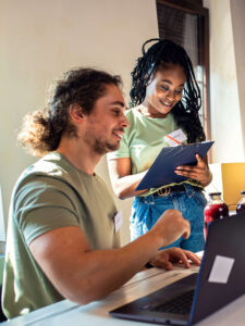 two community organization colleagues working together, with one typing on the laptop on the desk and the other taking notes