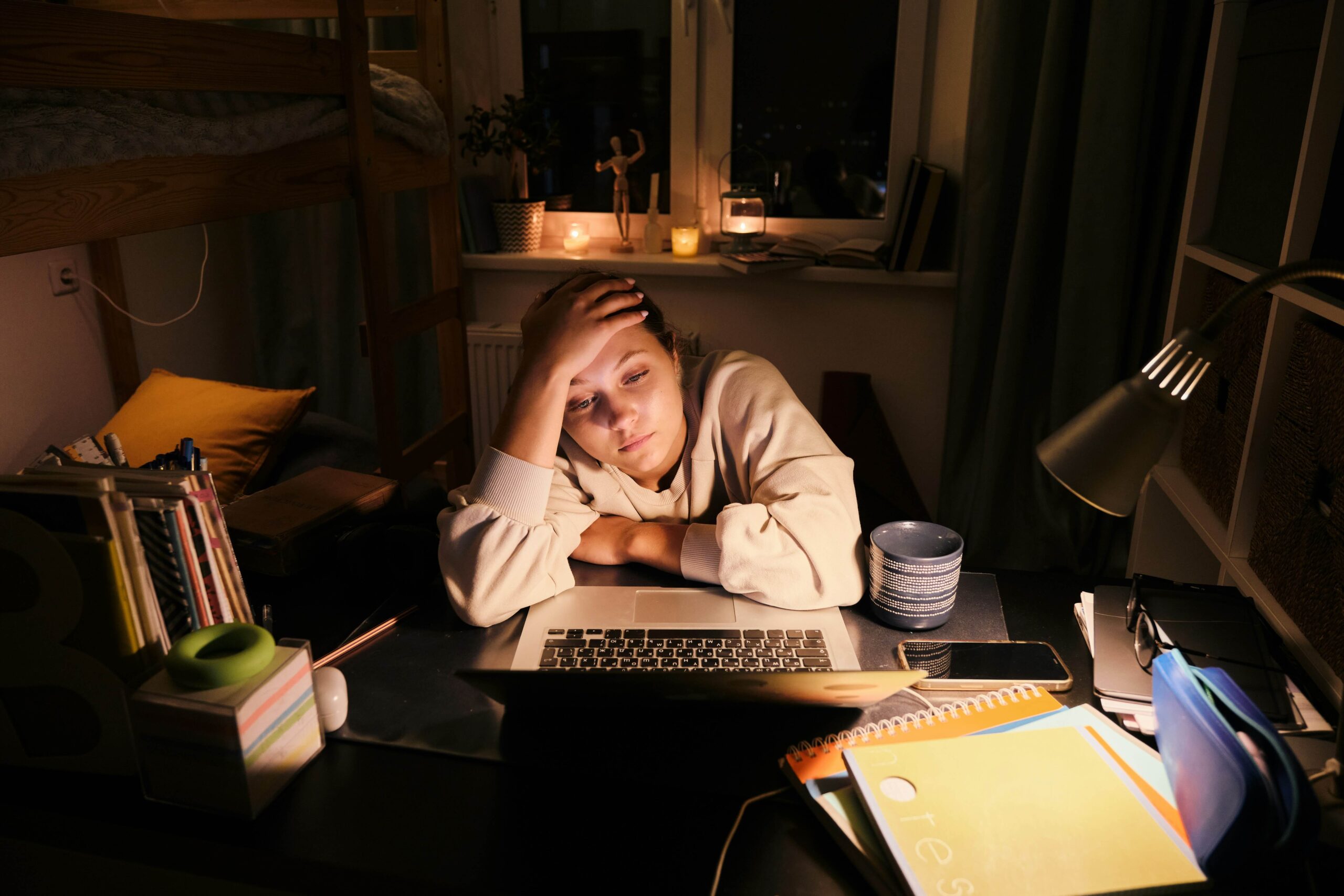 A young person sitting in front of their laptop in a dark room, holding their forehead and surrounded by clutter.