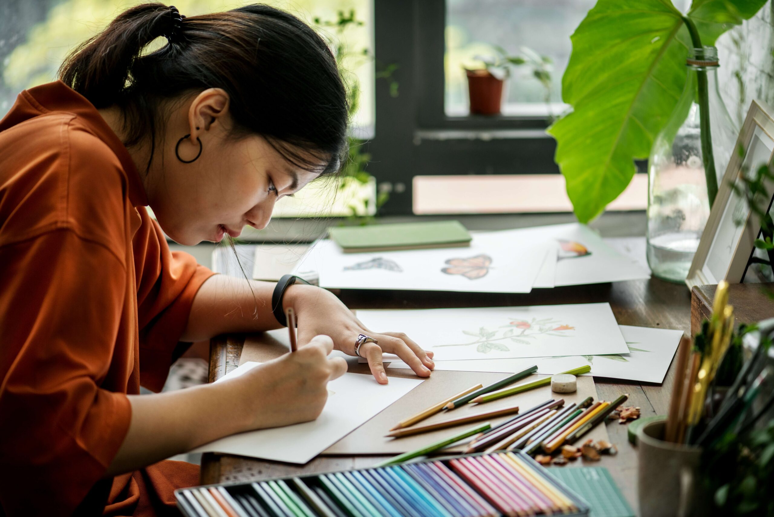 A young person wearing an orange shirt and black hoop earrings leans over a desk covered in colored pencils and artwork.