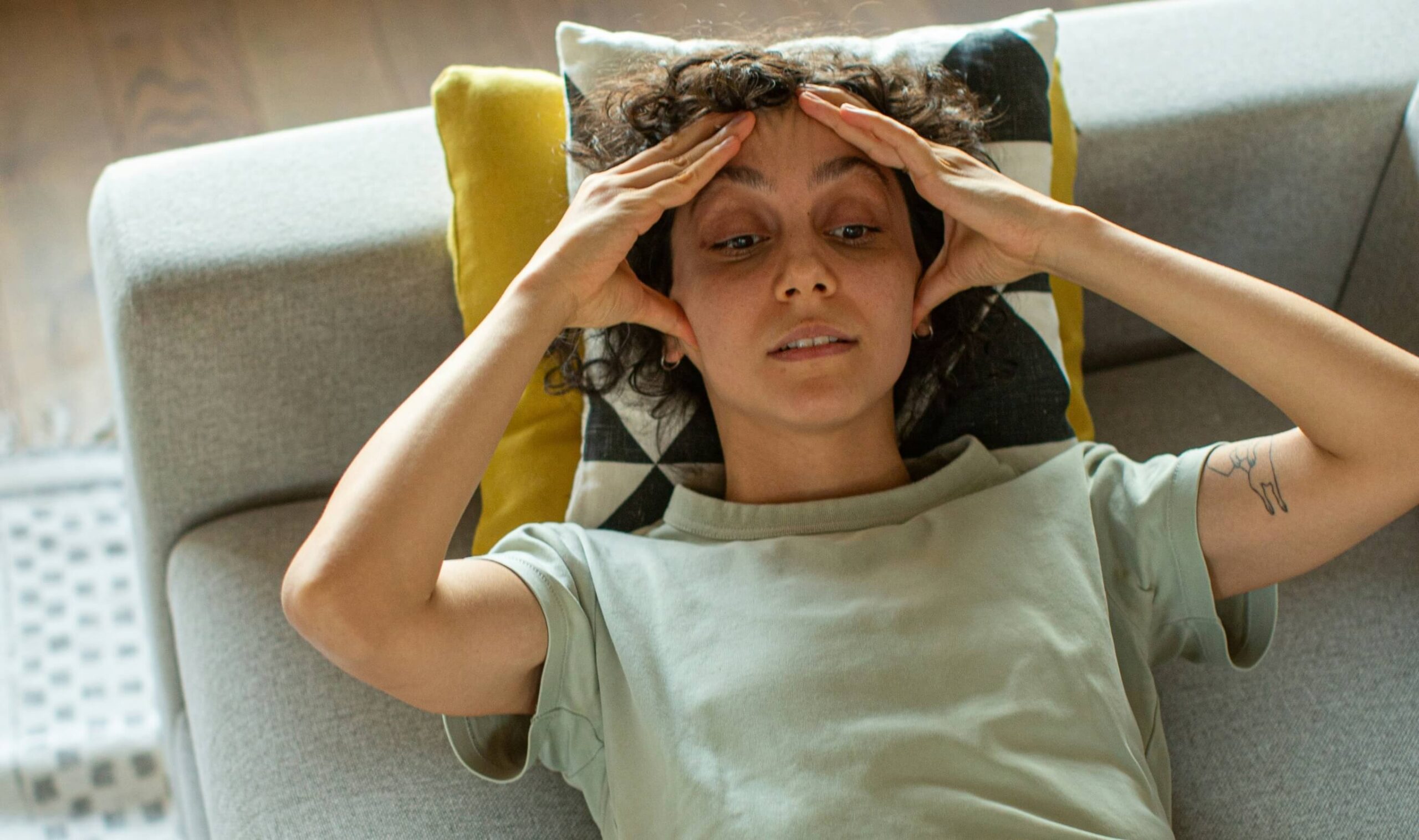 A young person lays on a gray couch with their hands cupping their forehead and a panicked expression on their face.