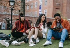 three students sitting on campus lawn talking. reading books and looking at a laptop
