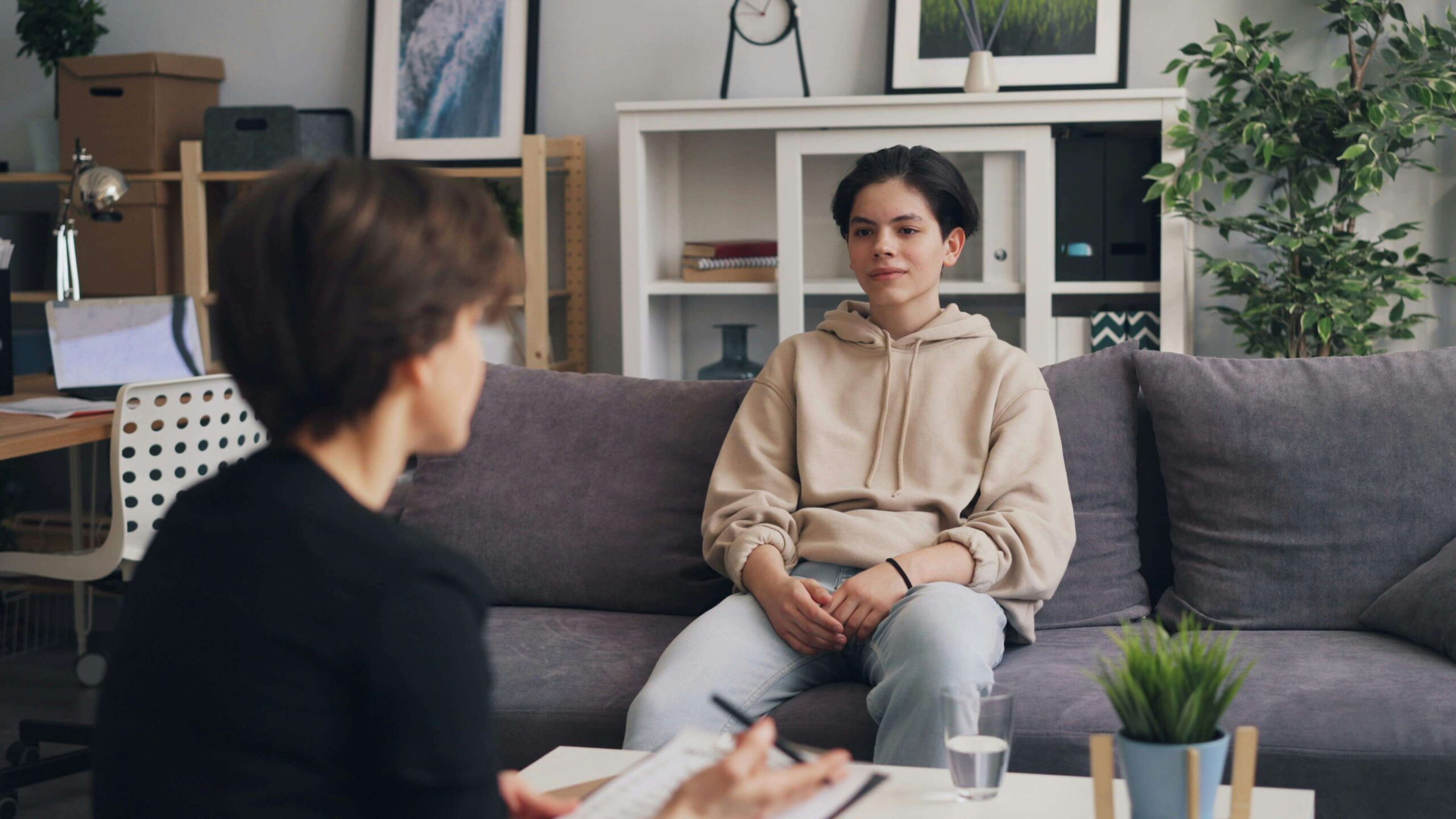 A young person sits on a gray couch in a therapist's office while their therapist listens and takes notes.